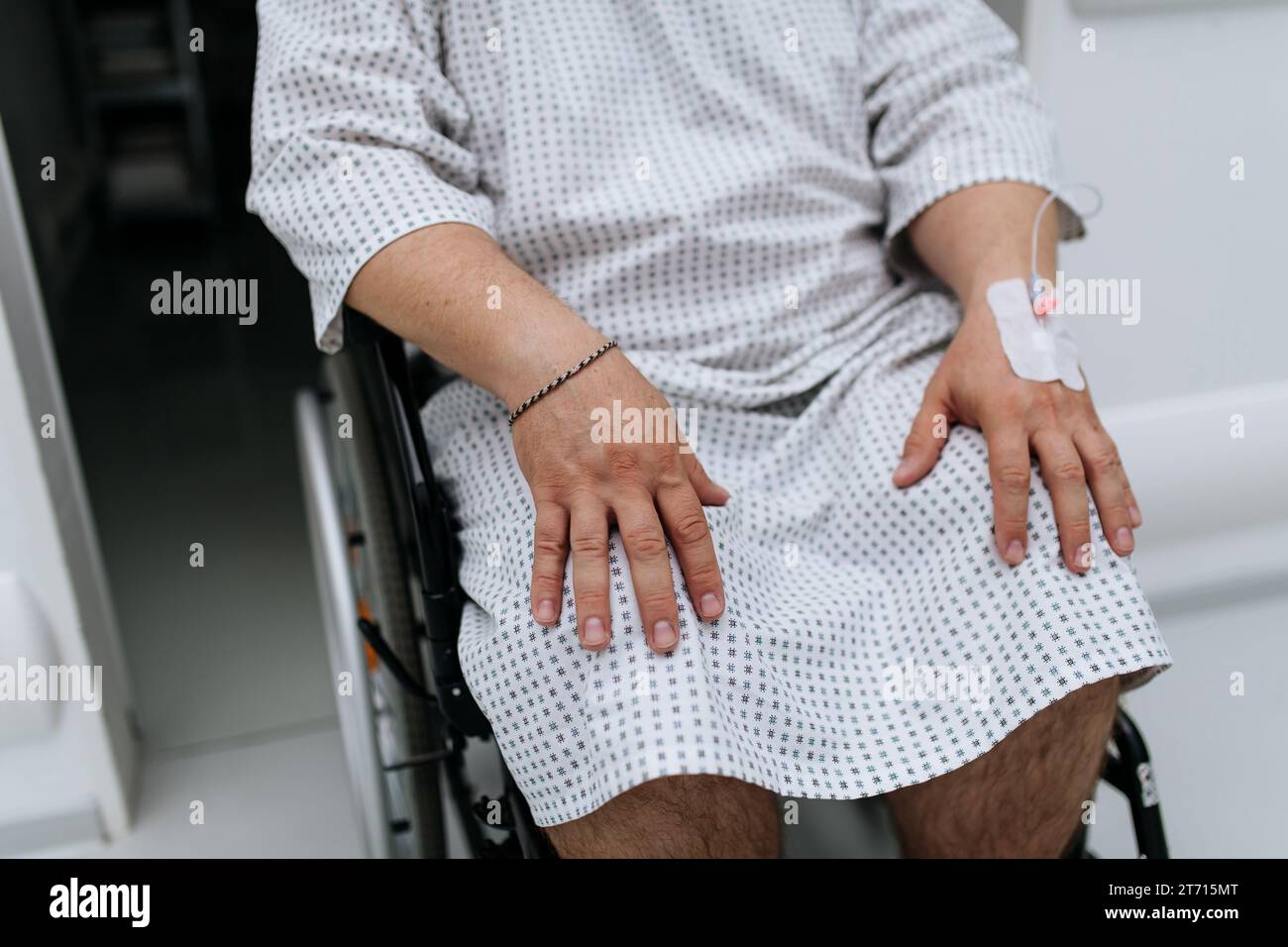 Primo piano di un uomo in sedia a rotelle con la cannula IV in mano. Paziente in sovrappeso in abito ospedaliero in attesa di visita medica in ospedale. Foto Stock