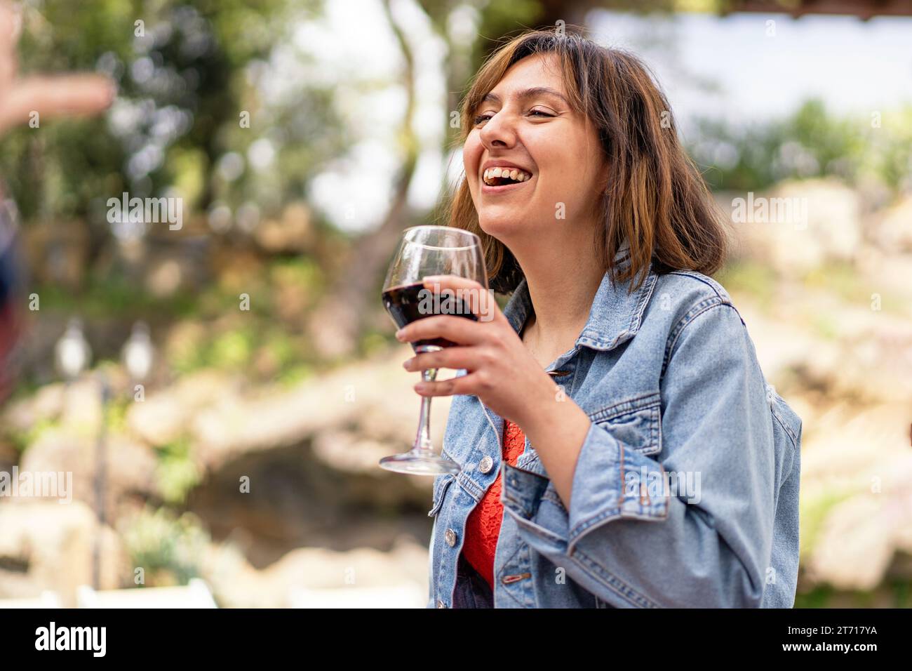 Una donna con una giacca in denim ride mentre si gusta un bicchiere di vino rosso, catturando un momento di pura gioia. Foto Stock