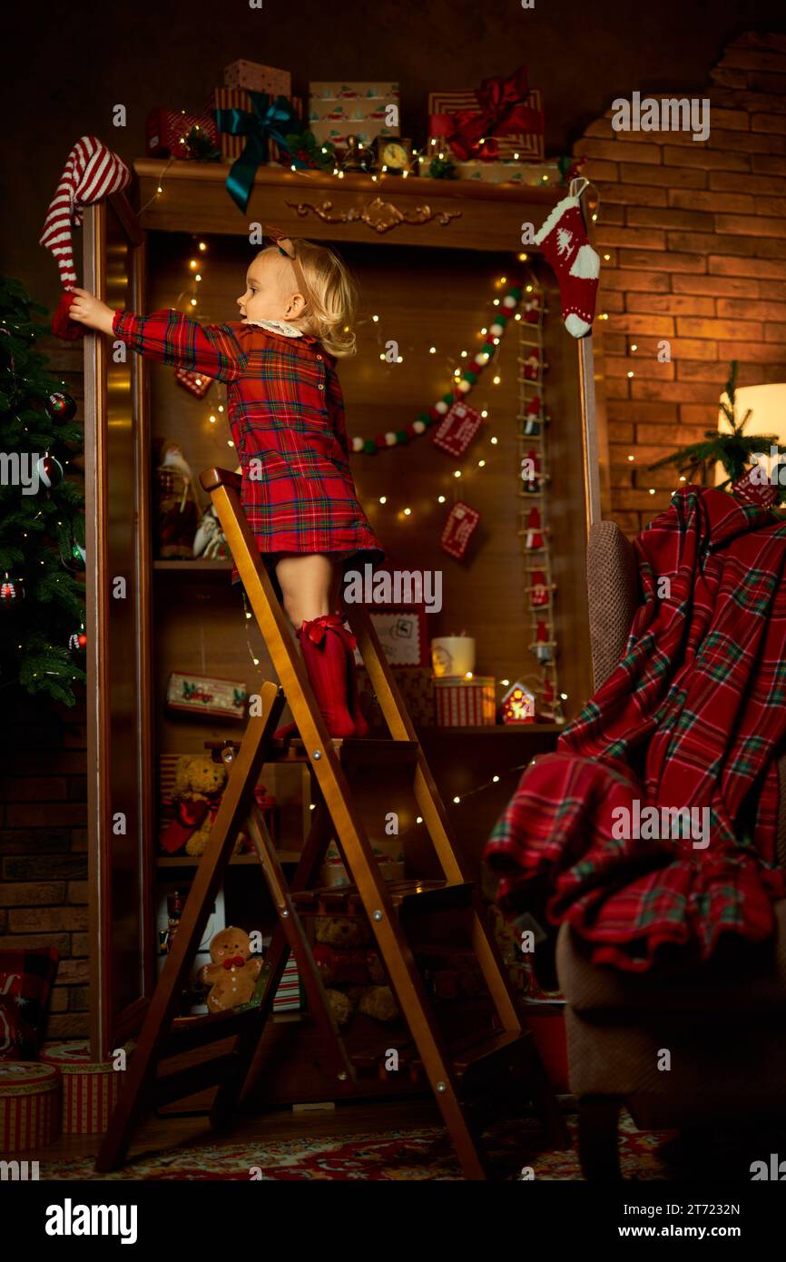 Buon Natale e buone feste. Una bambina con un abito a quadri sale le scale fino allo scaffale superiore di un armadio con regali. Atmosfera di magia, incisione Foto Stock