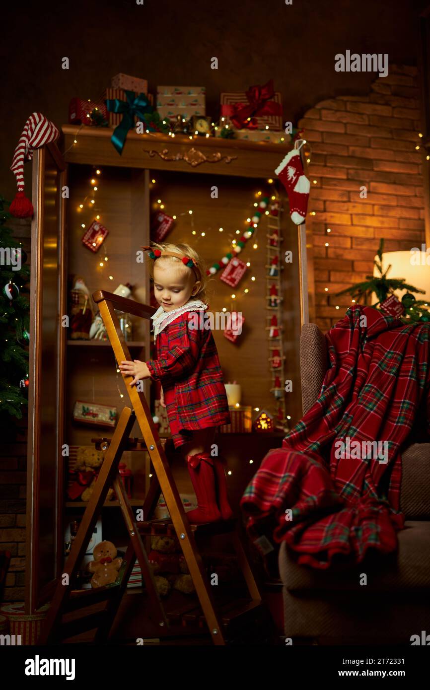 Buon Natale e buone feste. Una bambina con un abito a quadri sale le scale fino allo scaffale superiore di un armadio con regali. Atmosfera di magia, incisione Foto Stock