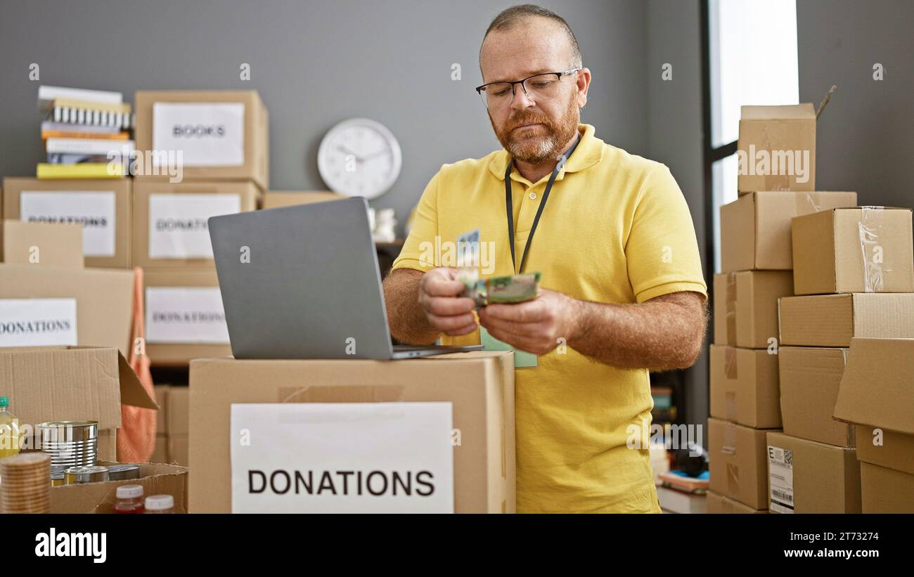 Bell'uomo caucasico barbuto che si offre volontario in un centro di beneficenza, contando seriamente i dollari australiani su un computer portatile tra scatole di donazioni Foto Stock