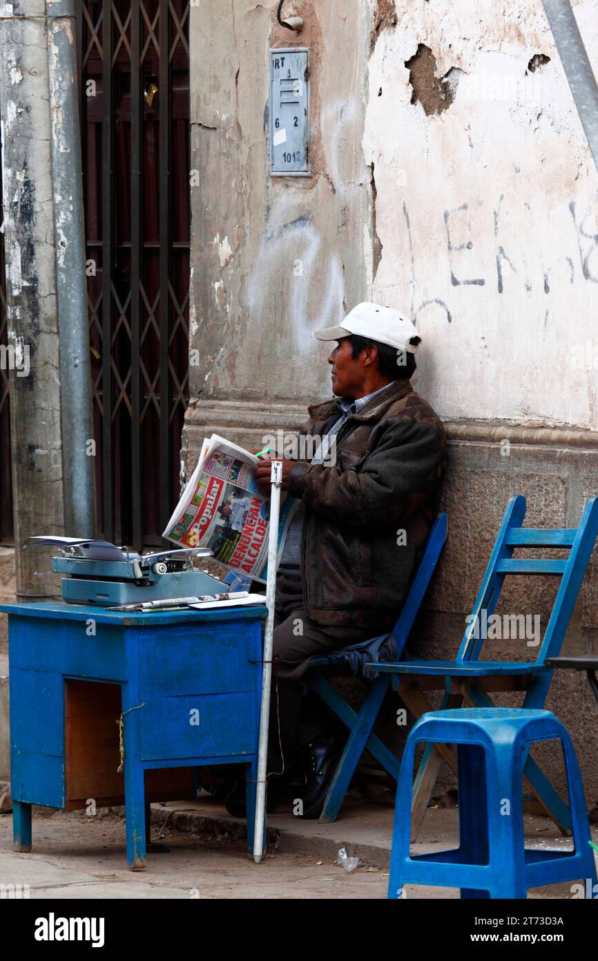 Un impiegato informale di ufficio di strada legge un giornale in attesa di affari, Puno, Perù. Molti di questi lavoratori in America Latina offrono i loro servizi compilando tutti i tipi di moduli, digitando lettere e affari simili a causa del gran numero di persone che non sanno leggere o scrivere. È comune vederne molti al di fuori del governo (locale), delle tasse e di altri uffici in cui le persone vanno a fare i documenti necessari. Foto Stock