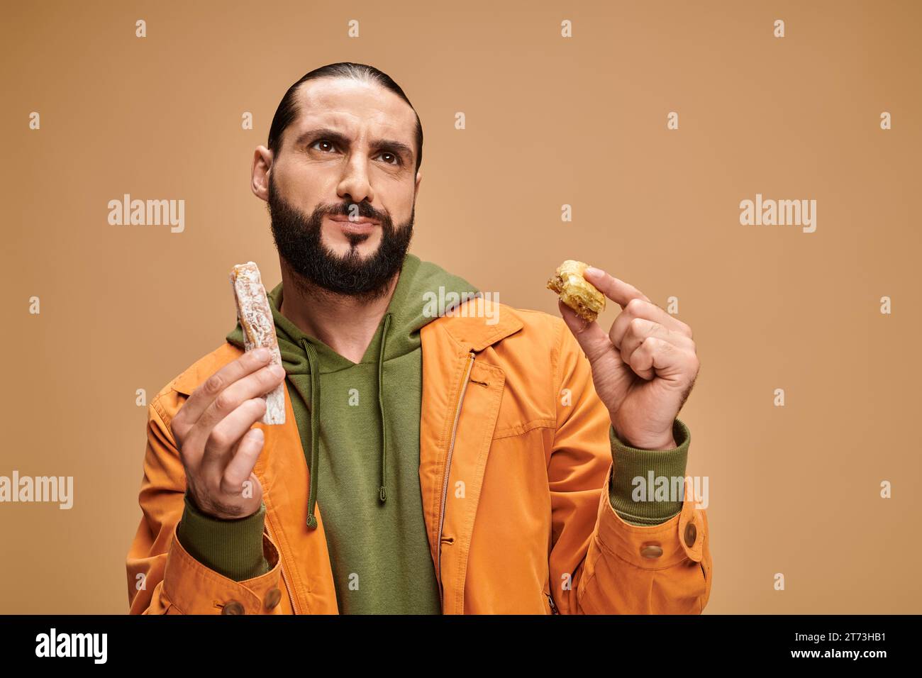 un uomo barbuto confuso che regge baklava e cevizli sucuk sullo sfondo beige, delizie turche Foto Stock