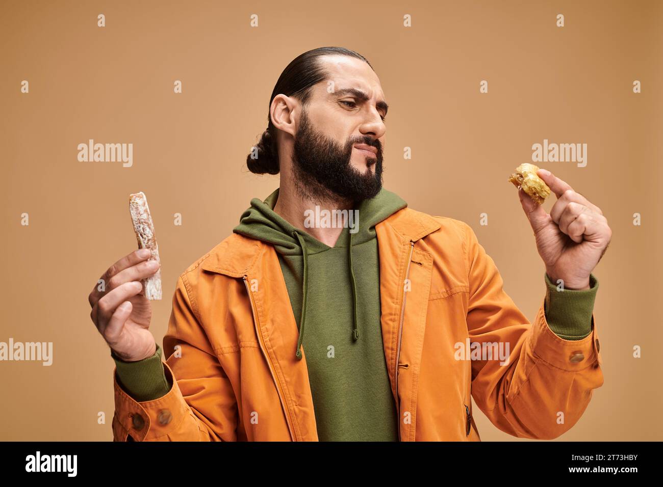 un uomo barbuto arabo confuso che regge baklava e cevizli sucuk sullo sfondo beige, delizie turche Foto Stock