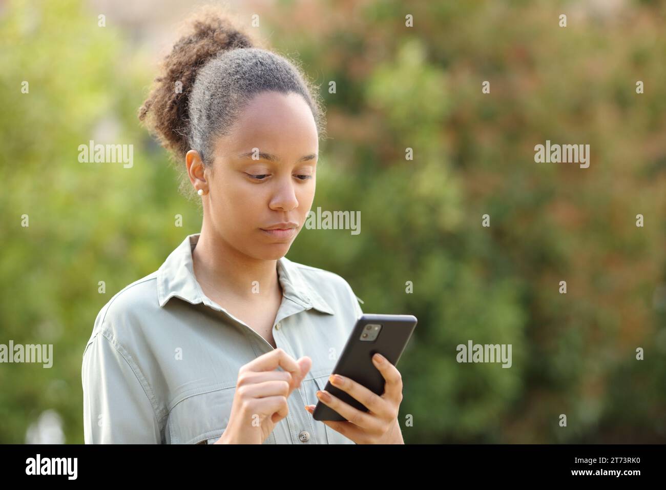 Una donna nera seria che cammina in un parco, controlla il telefono Foto Stock