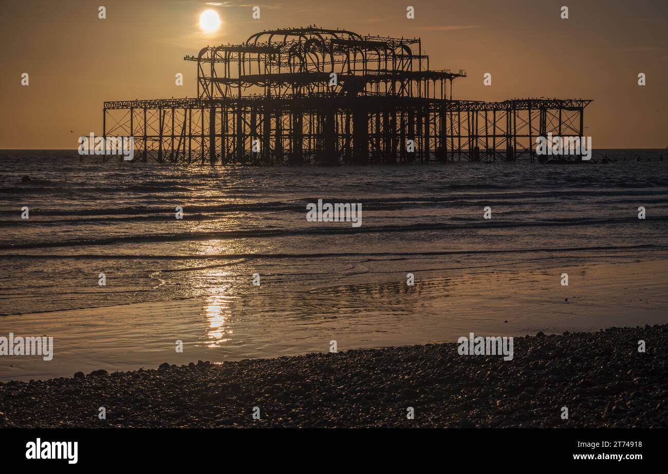 Il sole tramonta dietro le rovine del vecchio molo occidentale sul lungomare di Brighton, Sussex, Inghilterra, Regno Unito Foto Stock