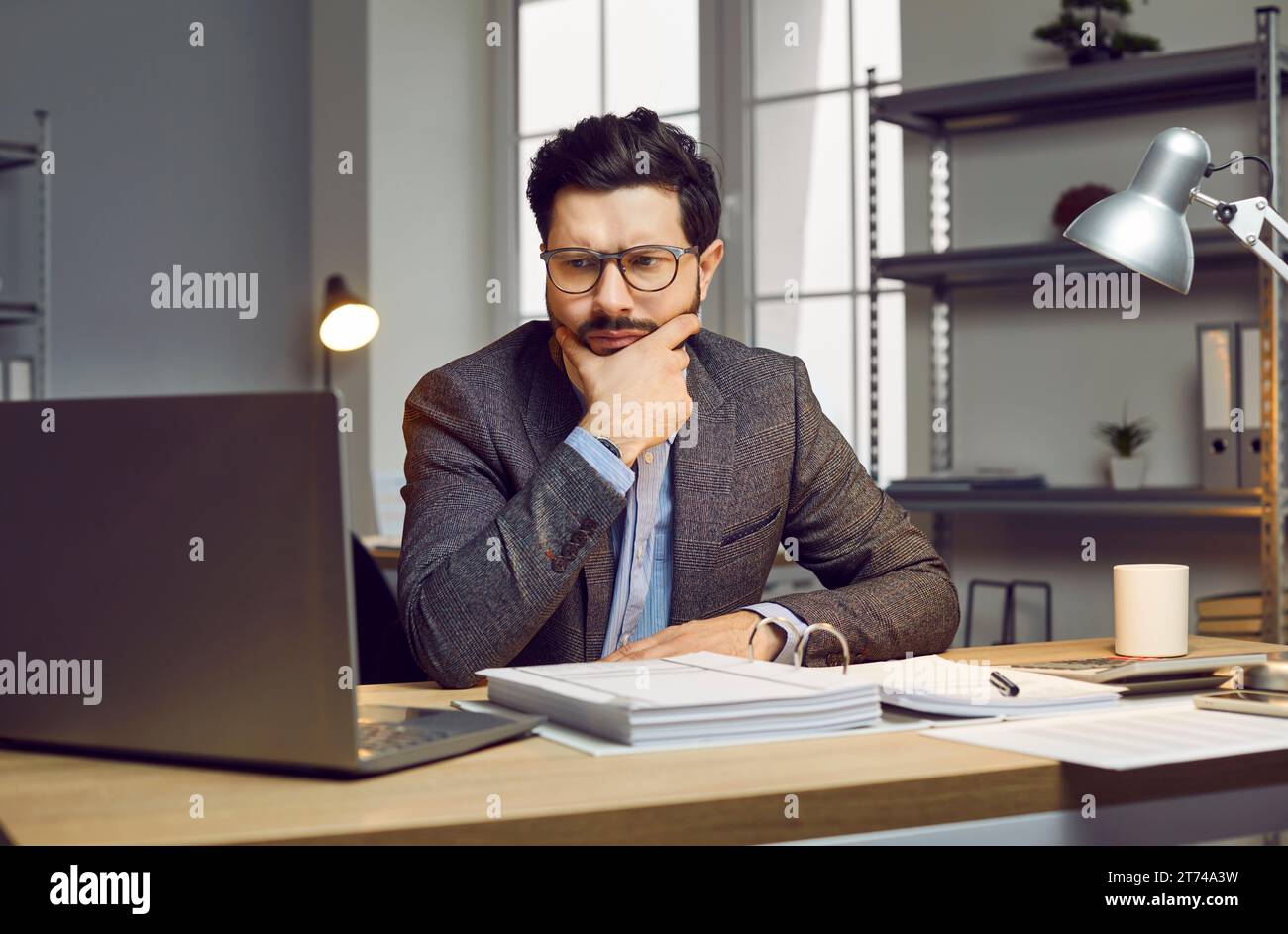 Ritratto di un giovane uomo d'affari serio, premuroso, attraente con occhiali, seduto sul posto di lavoro e guardando lo schermo del suo notebook. Foto Stock