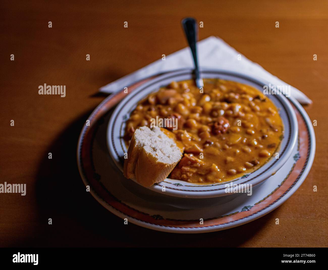 TIPICO FABADA SPAGNOLO IN UN PIATTO, CON UN PEZZO DI PANE E UN CUCCHIAIO. TUTTO È SU UN TAVOLO DI LEGNO CON UN TOVAGLIOLO Foto Stock