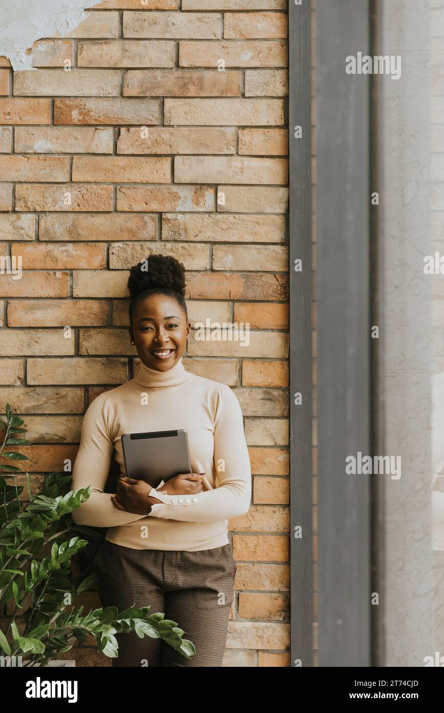 Una bella giovane donna d'affari afroamericana con un tablet digitale in piedi accanto al muro in stile industriale Foto Stock
