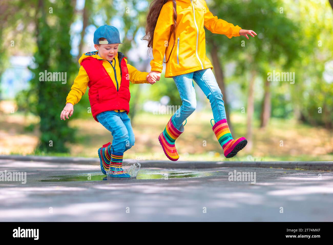 Ragazza e ragazzo felici con ombrello e stivali da pioggia colorati in gomma giocando all'aperto e saltando in pozza piovosa Foto Stock
