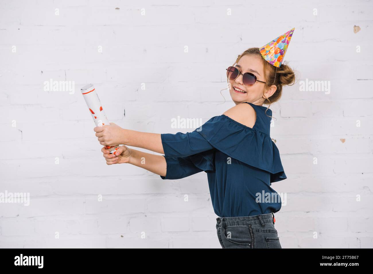 Ragazza adolescente sorridente che lascia fuori la festa popper in piedi davanti al muro bianco Foto Stock