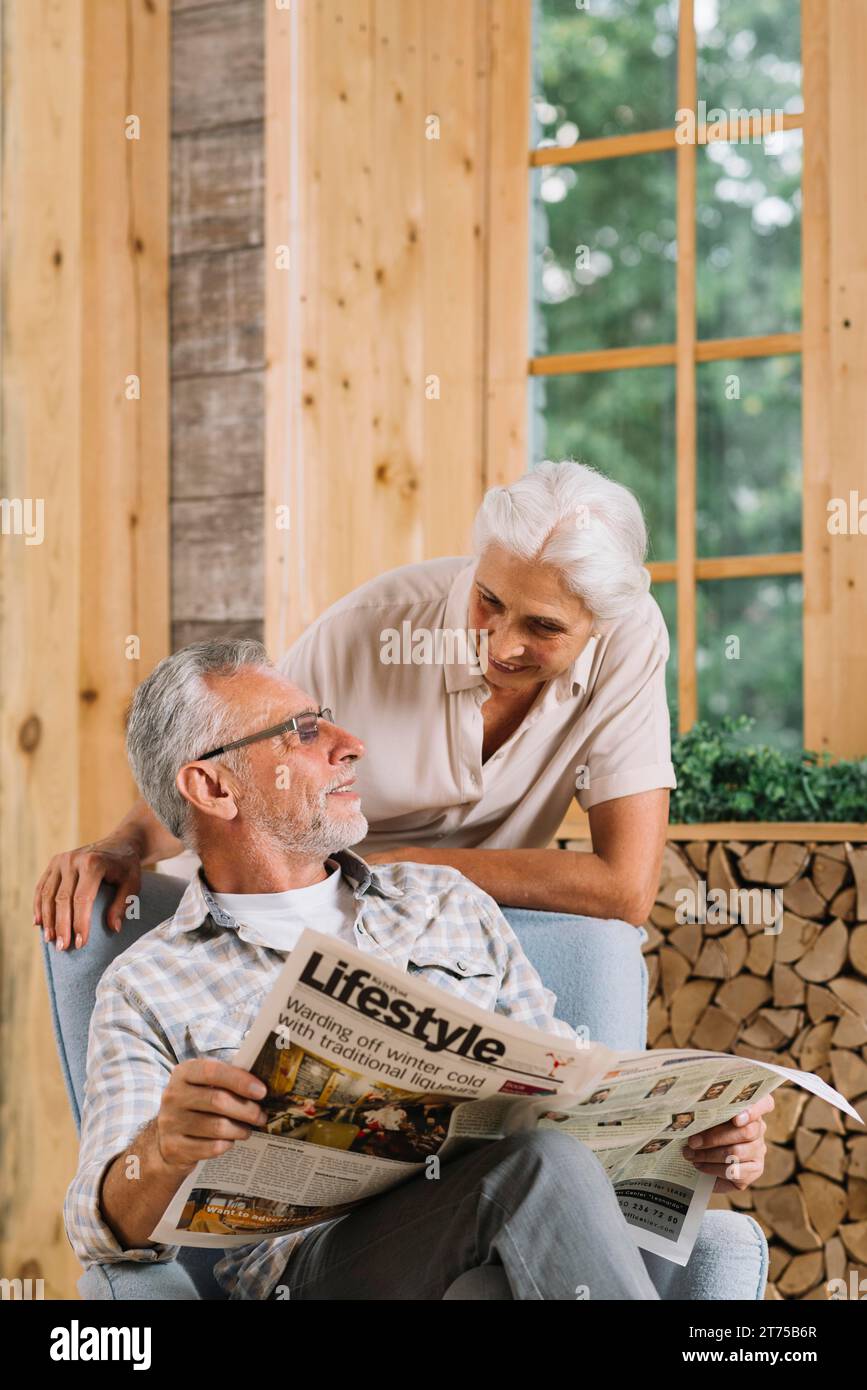 Donna anziana sorridente che guarda suo marito seduto su una sedia che tiene un giornale Foto Stock