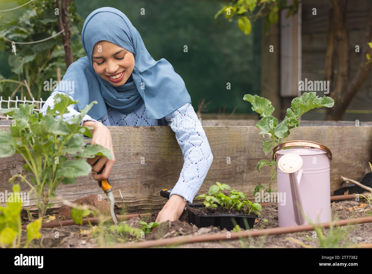 Felice donna birazziale in hijab che piantina in un giardino soleggiato Foto Stock