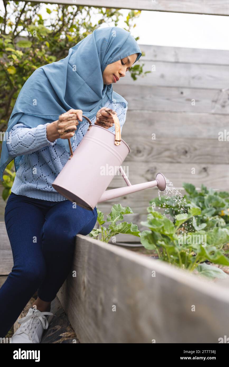 Donna birazziale in hijab che annaffiano piante in giardino soleggiato Foto Stock