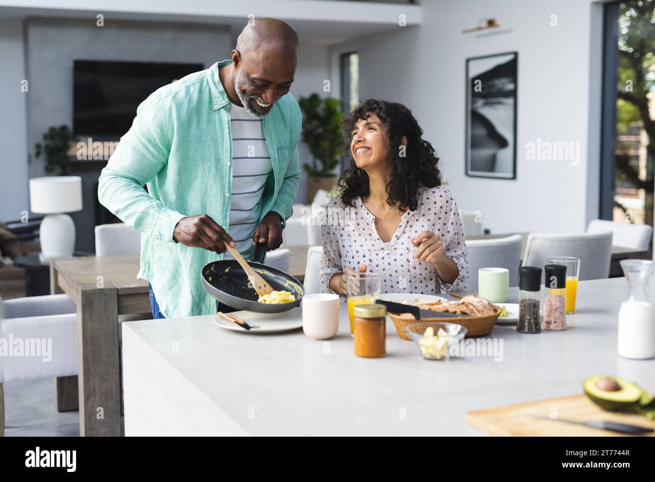 Coppia matura e diversificata che serve uova strapazzate e che fa colazione nella soleggiata cucina, spazio fotocopie Foto Stock
