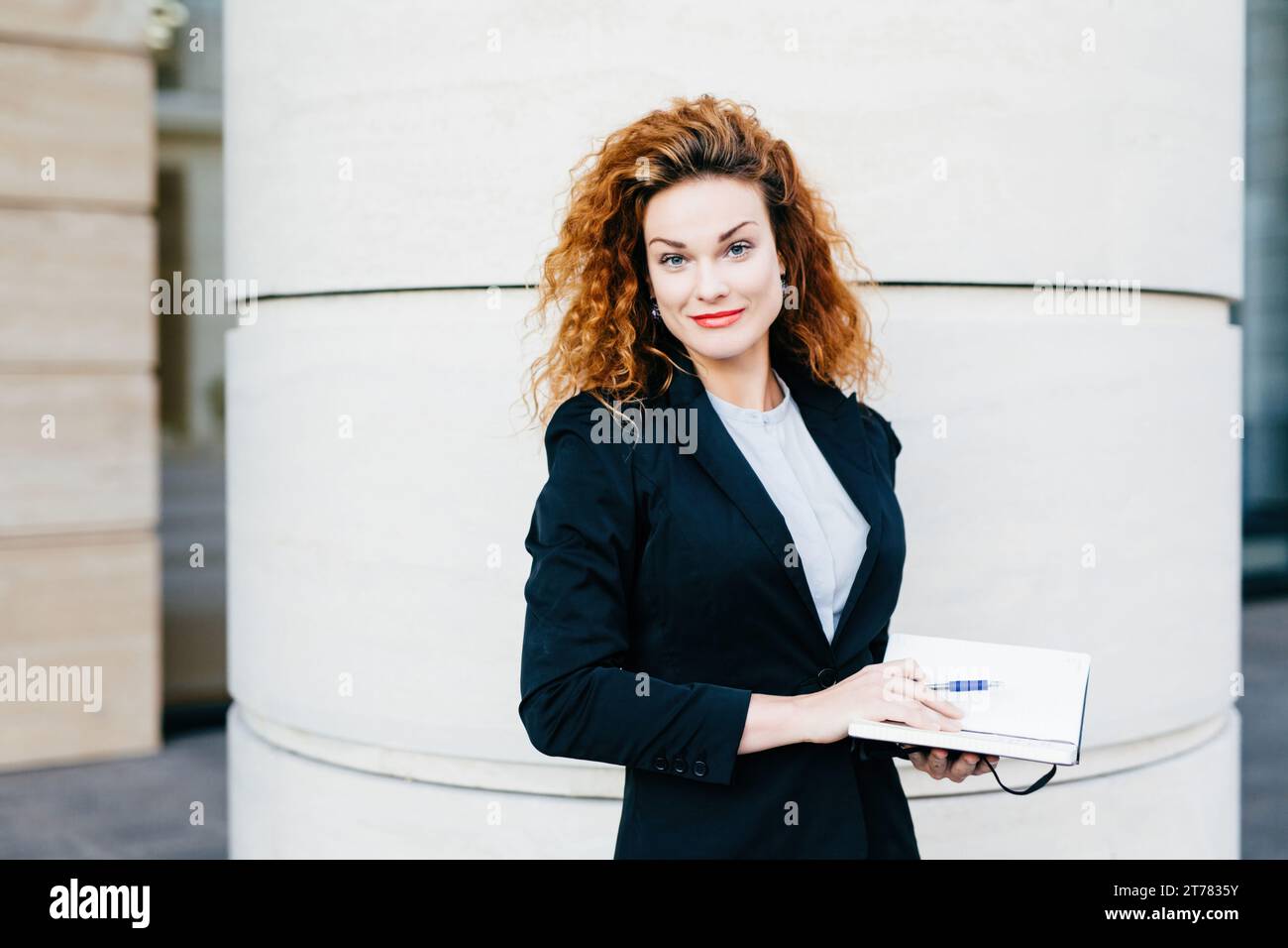 Una donna d'affari dai capelli ricci e splendenti, che stringe un notebook, si trova davanti a una colonna, incarnando la sicurezza Foto Stock