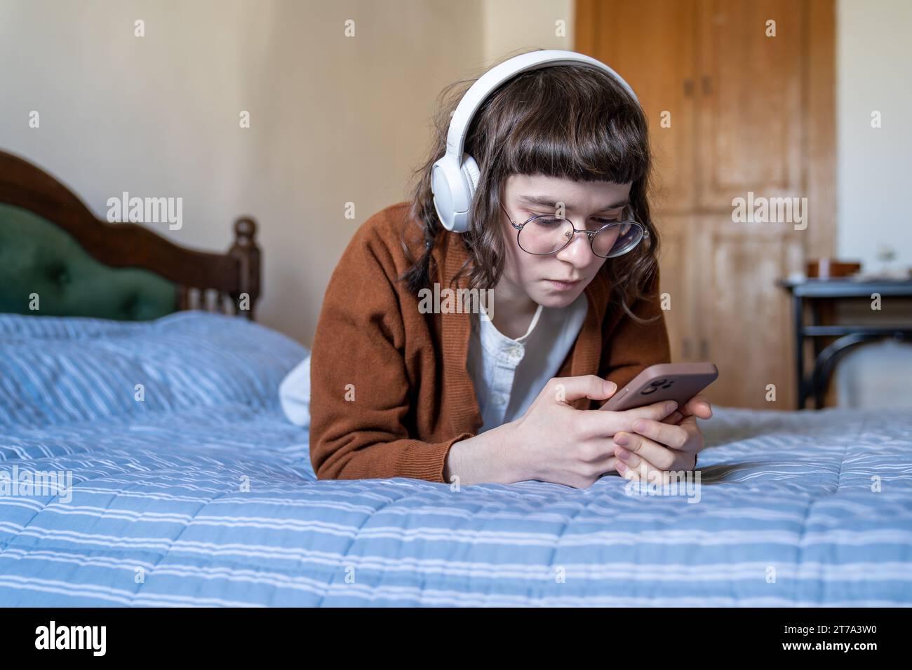 Ragazza adolescente seria sdraiata sul letto, guardando film, video educativi, ascoltando musica sullo smartphone Foto Stock