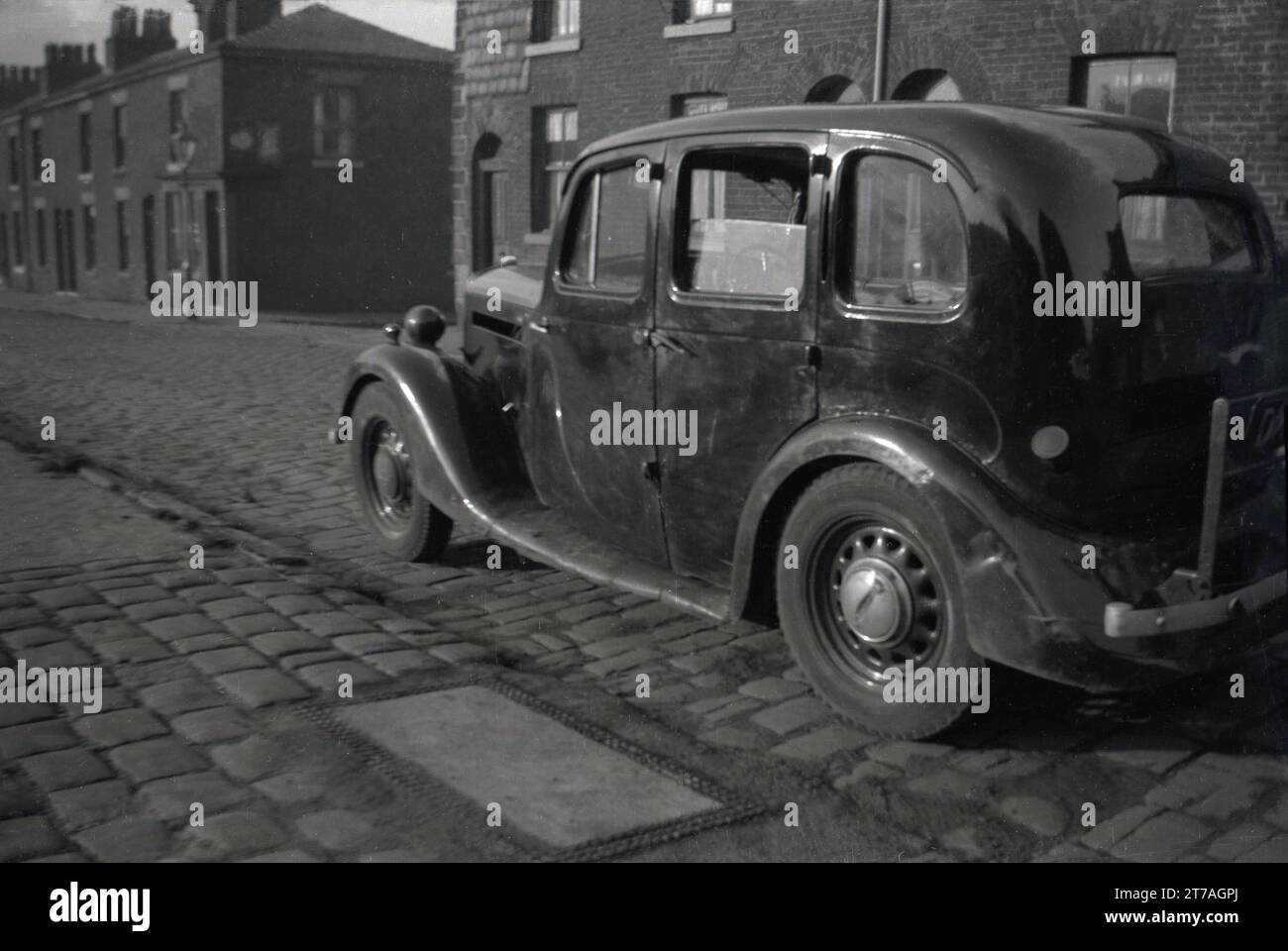 Anni '1950, storica, un'automobile di un decennio precedente, probabilmente degli anni '1940, parcheggiata su una strada acciottolata di piccole case vittoriane terrazzate, Oldham, Manchester, Inghilterra, Regno Unito. Foto Stock