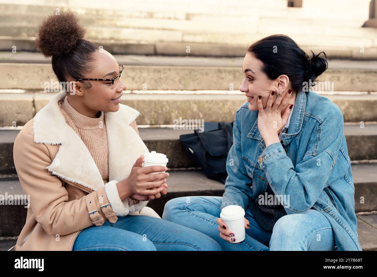 due studentesse in una giacca di denim parlano tra loro, al telefono, ridono, bevono caffè sulle scale in città. Foto Stock