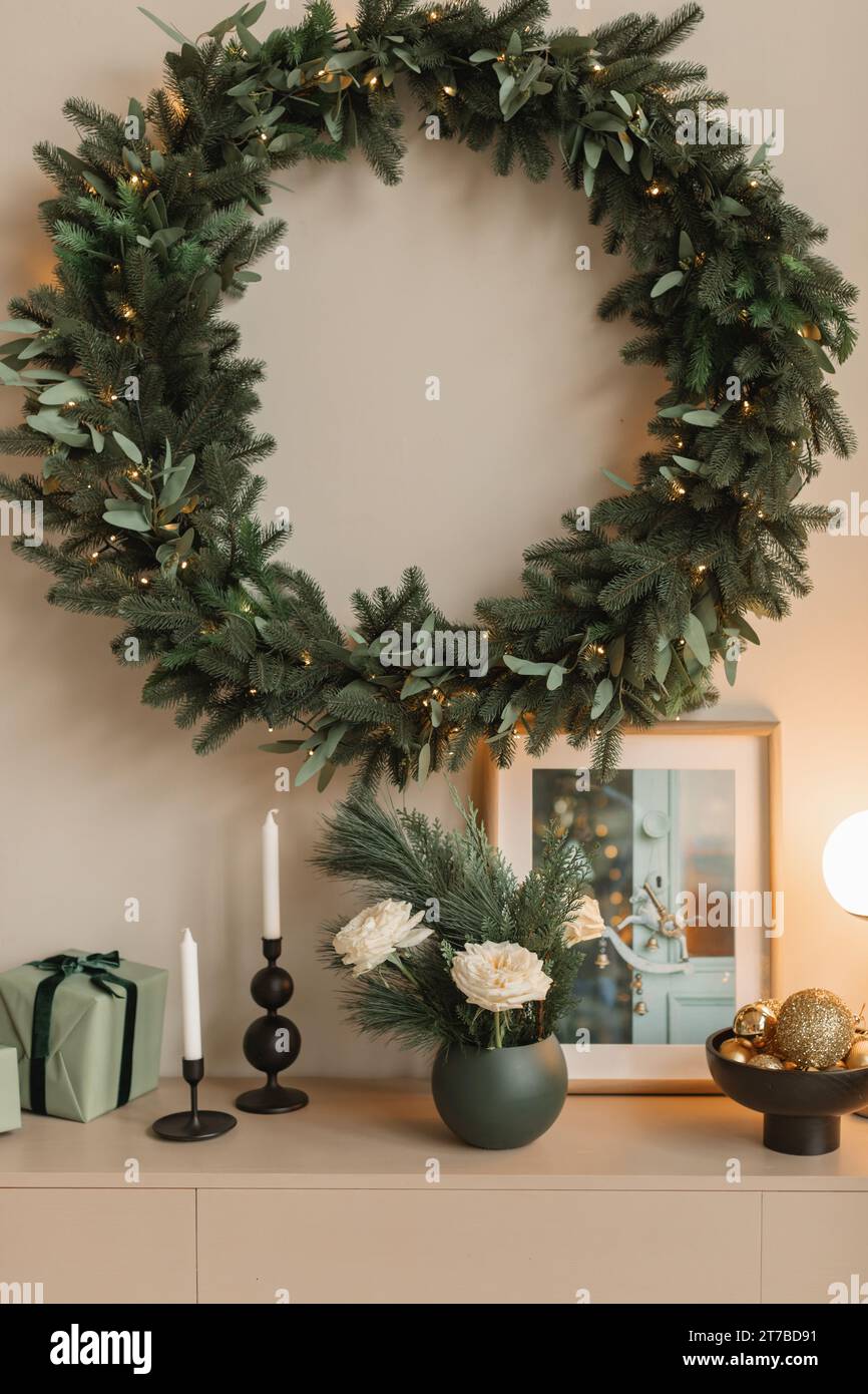 Corona di Natale illuminata appesa a una parete su una credenza con regali, candele, fiori, lampada e cornice Foto Stock