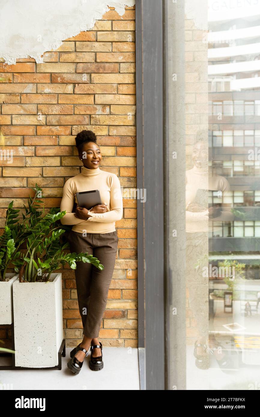 Una bella giovane donna d'affari afroamericana con un tablet digitale in piedi accanto al muro in stile industriale Foto Stock