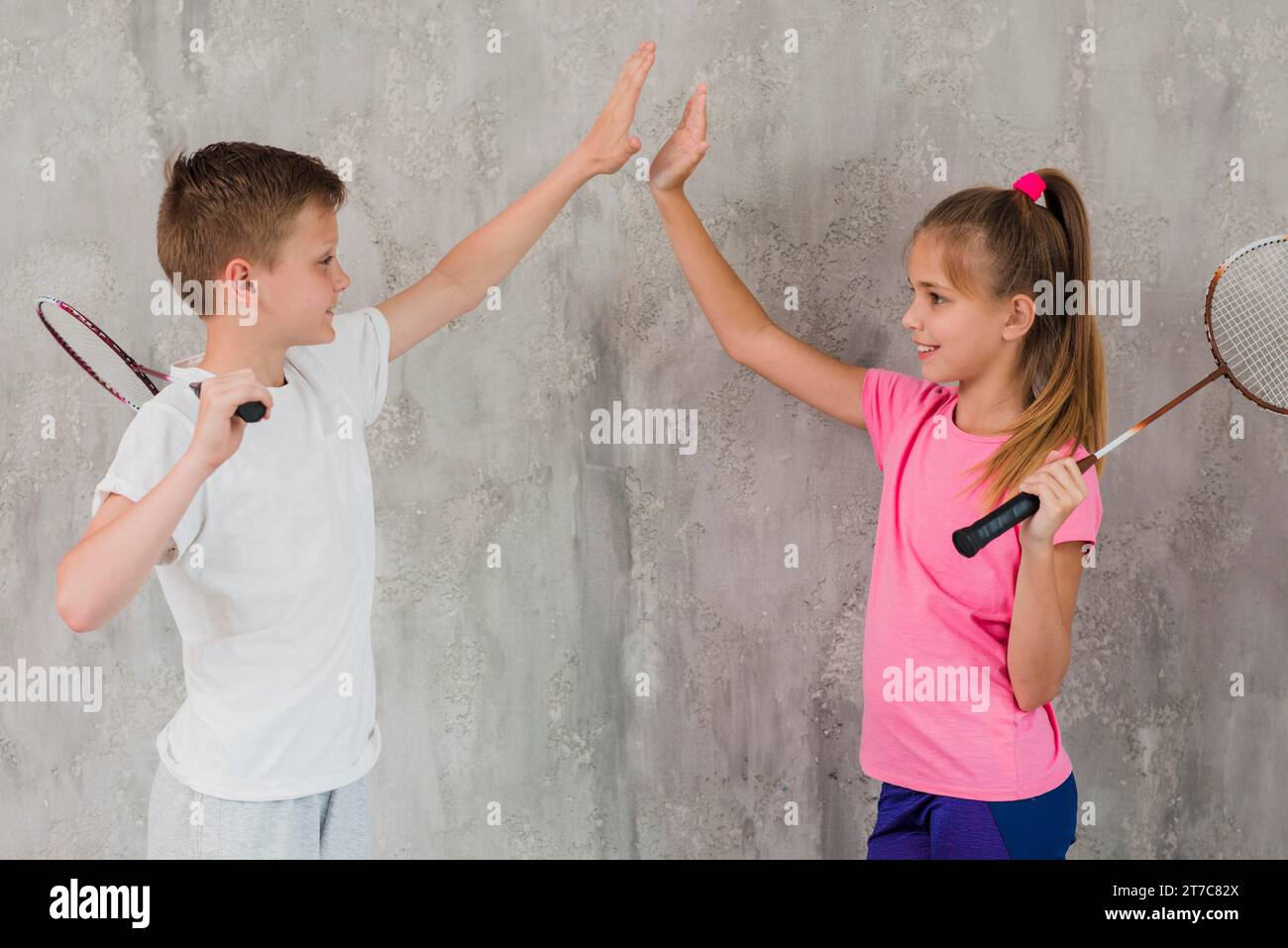 Bambina con vista laterale che tiene la racchetta e ne dà cinque in piedi contro il muro Foto Stock