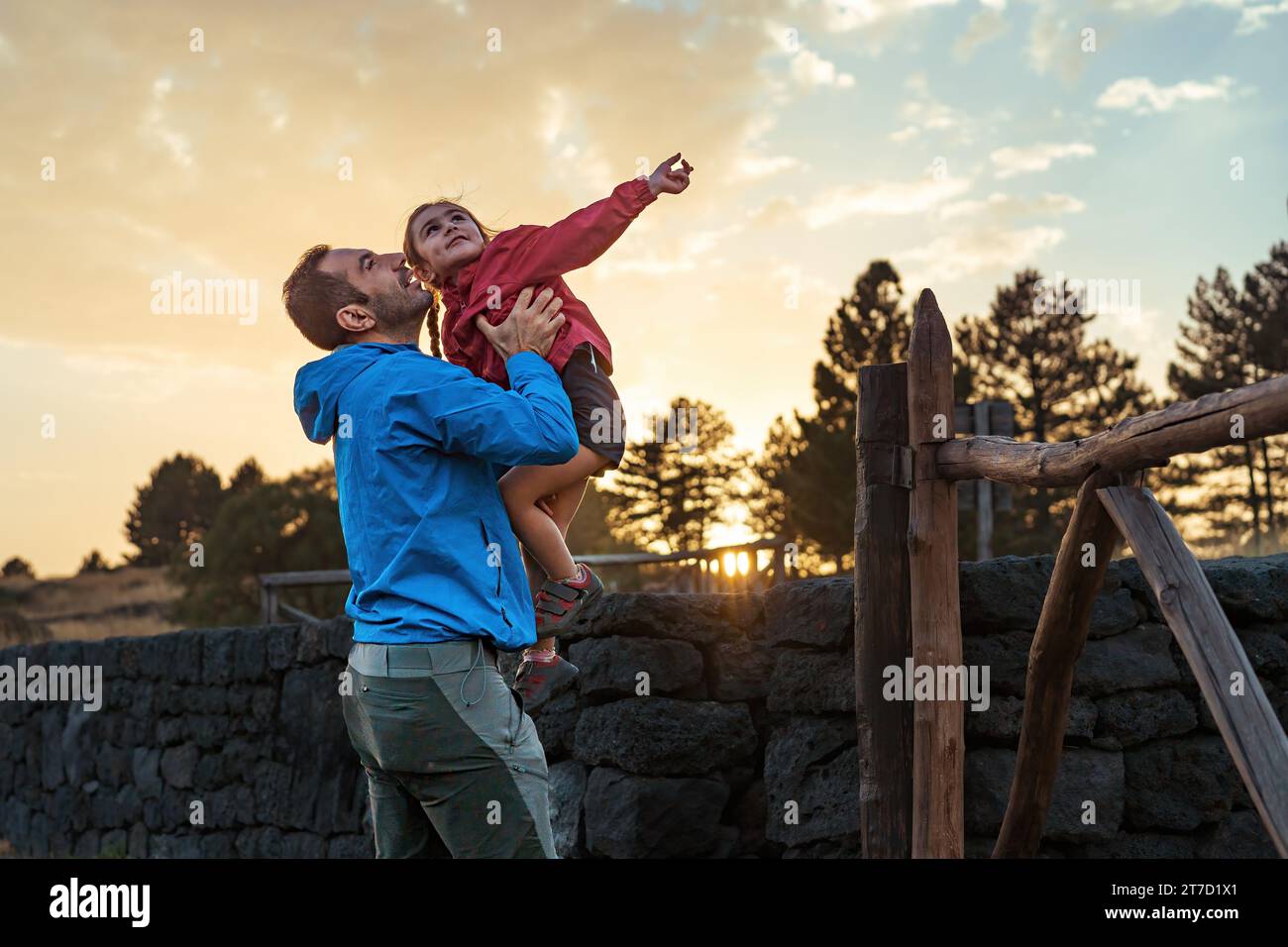 Padre allegramente solleva sua figlia mentre si gode un tramonto insieme, catturando un momento gioioso di legame con la famiglia. Foto Stock