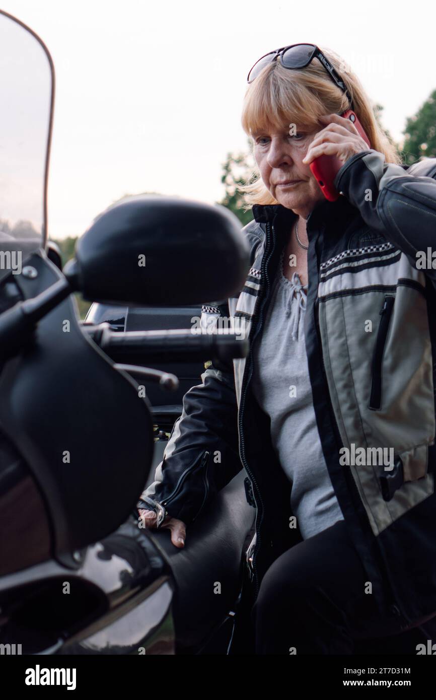 Ritratto verticale di una motociclista bionda matura che parla al telefono cellulare mentre è seduta sulla sua moto. Comunicazione durante i viaggi. Hobby - moto t Foto Stock