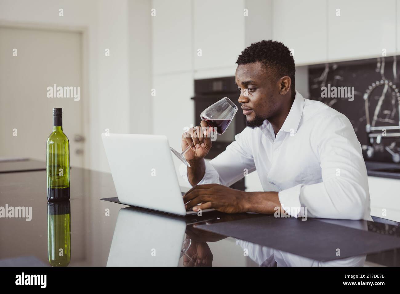 Un uomo d'affari afro-nero solitario si sente stanco stanco seduto a lavorare online e beve vino da solo a casa Foto Stock