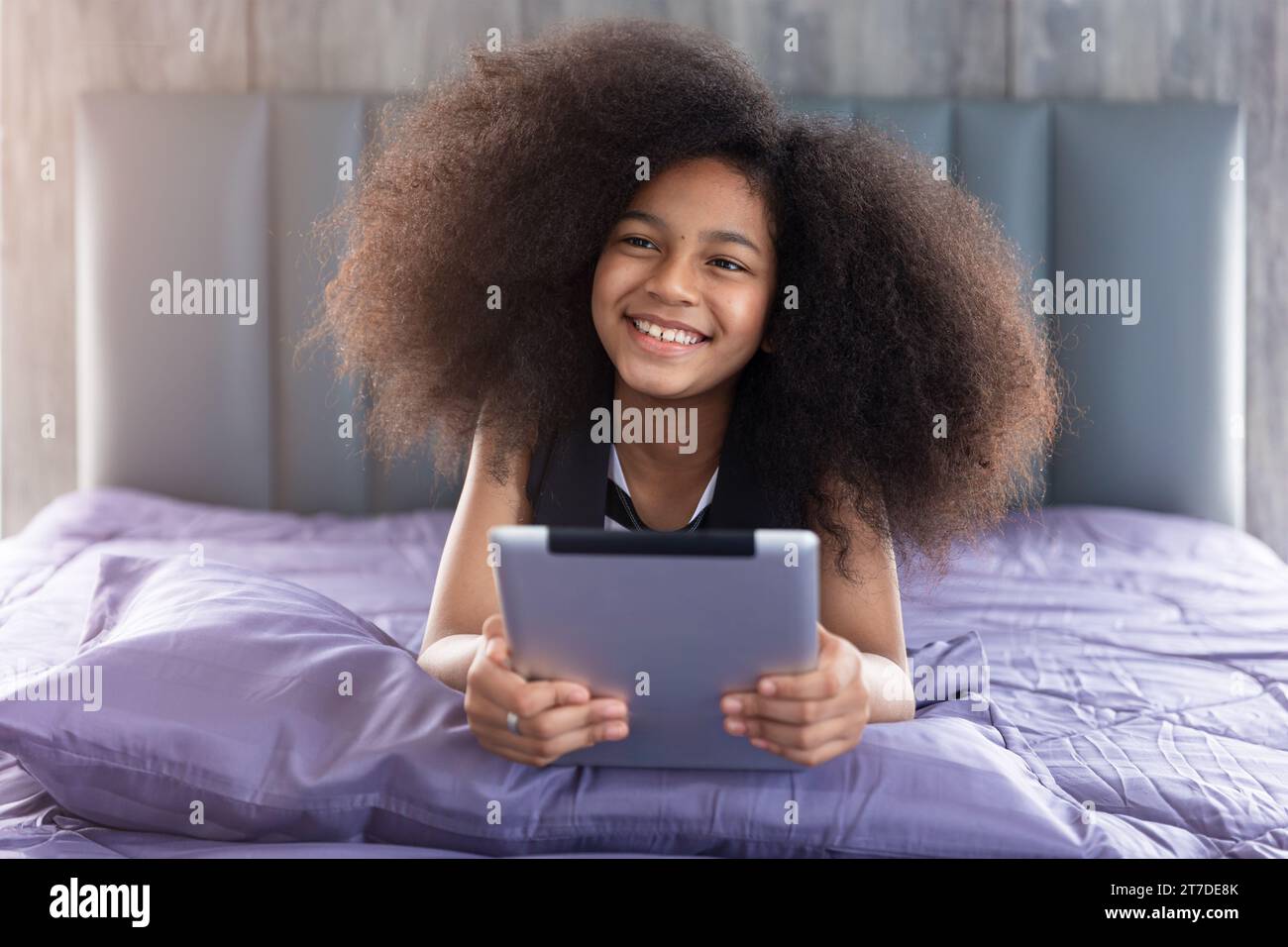 La bambina afro nera allegra e sorridente si trovava a letto con un tablet e un'istruzione di apprendimento a casa con un dispositivo digitale tecnologico. Foto Stock