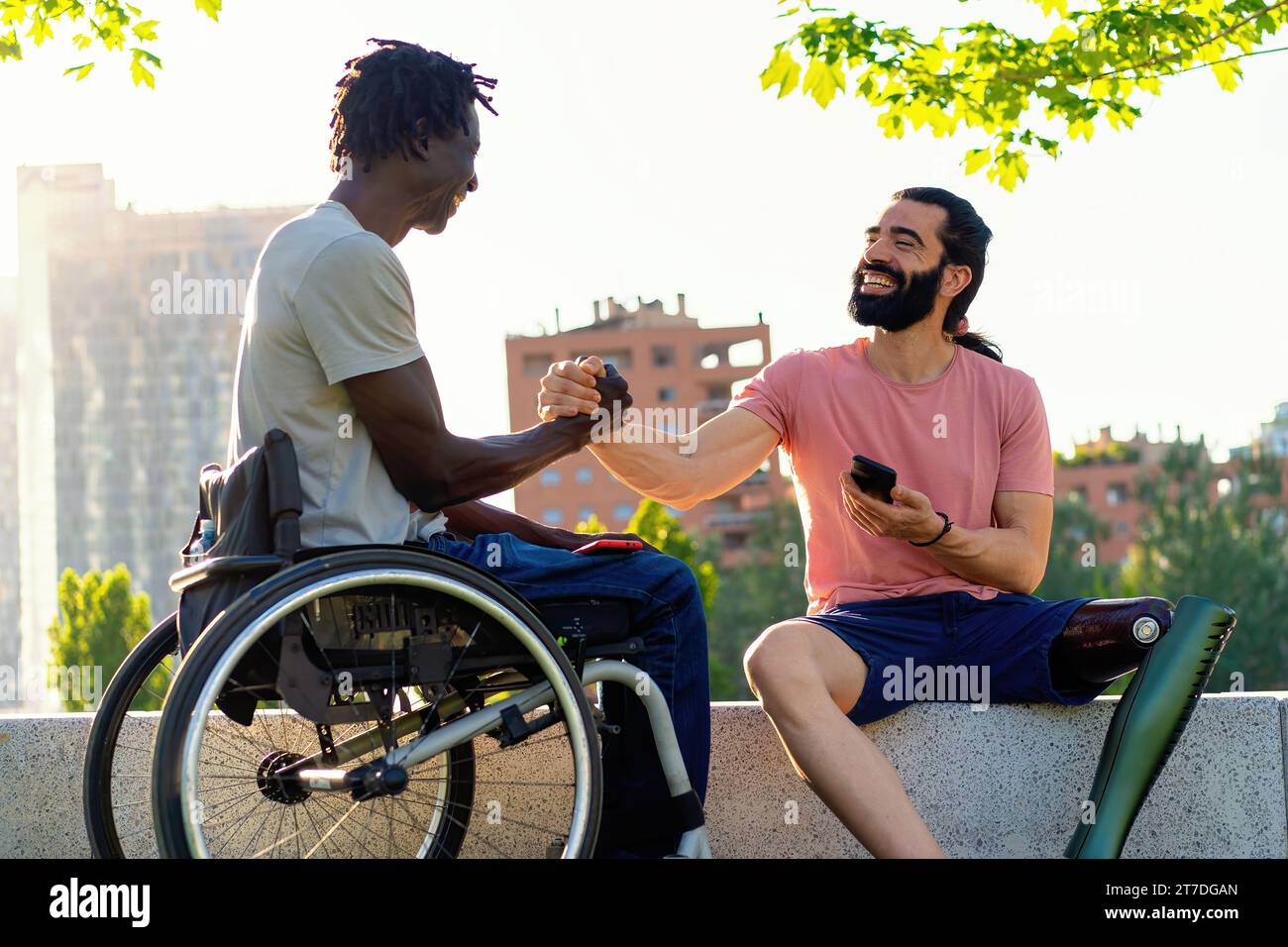 Due uomini, uno con una gamba protesica e uno in sedia a rotelle, condividono una stretta di mano in un ambiente urbano all'aperto. Foto Stock