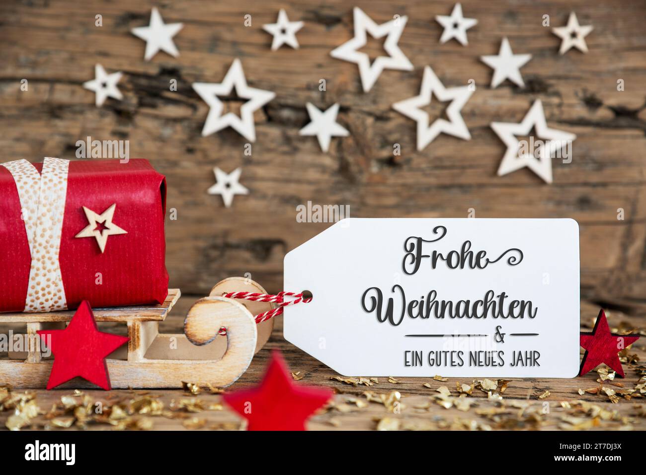 Sled in legno con regalo rosso e sfondo natalizio rustico con etichetta con testo tedesco Frohe Weihnachten und Ein Gutes Neues Jahr, che significa Merry CH Foto Stock
