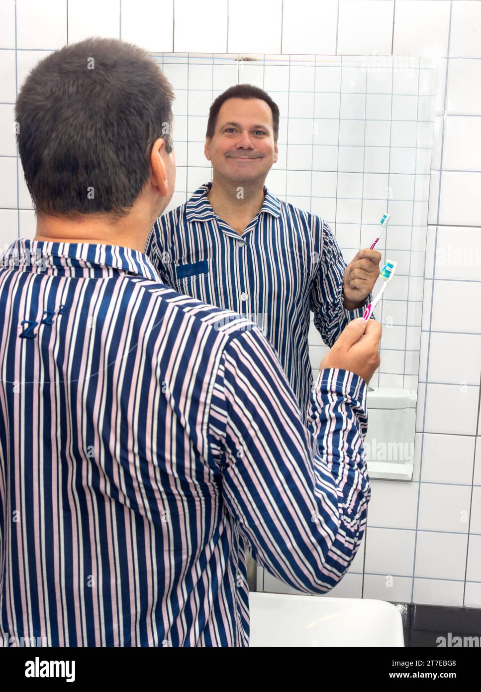 Un uomo sorridente in pigiama tiene uno spazzolino da denti e guarda nello specchio del bagno Foto Stock