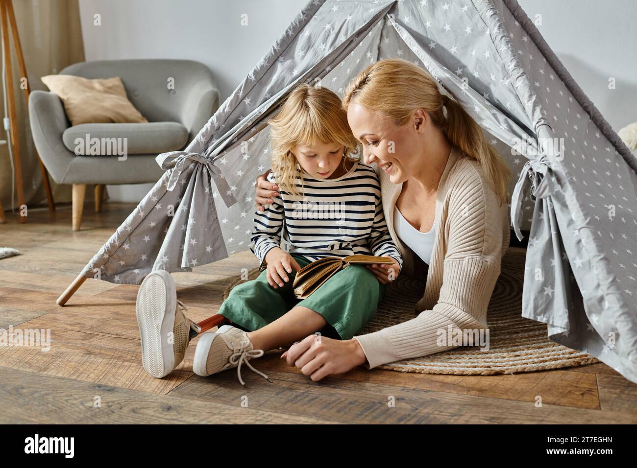 una ragazza carina con una gamba protesica che legge un libro e siede con una madre felice in una tenda da da gioco a casa Foto Stock