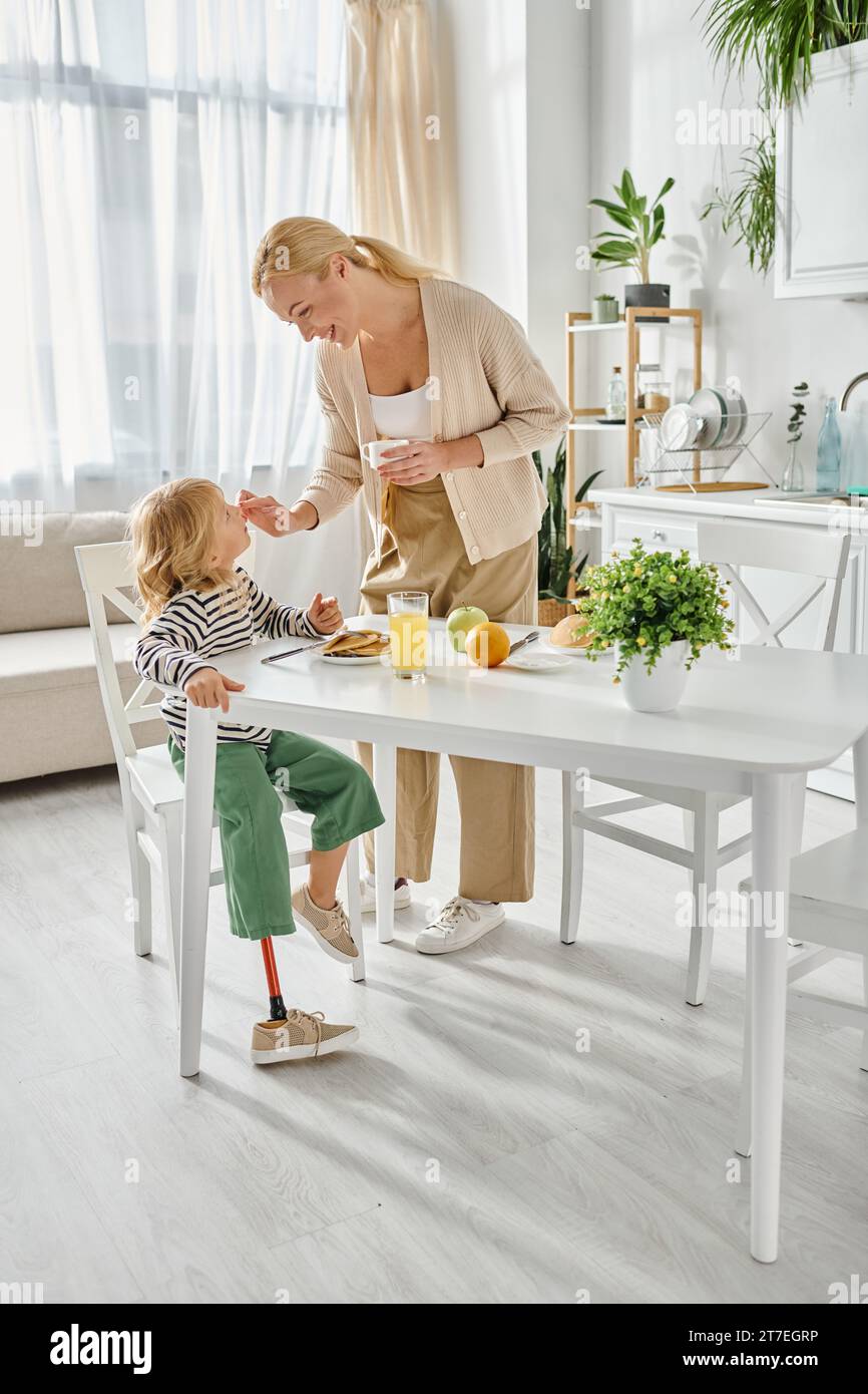 madre felice in piedi vicino a una figlia con protesi alla gamba che fa colazione in cucina, adattiva Foto Stock