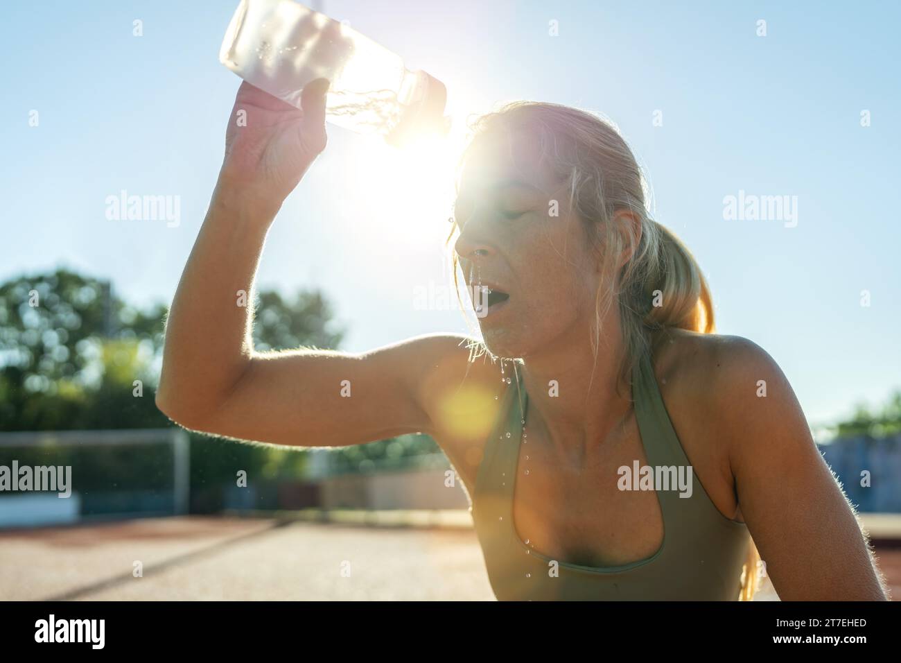 Donna che versa acqua sul viso dopo l'allenamento, luce solare, bottiglia sportiva, rinfresco Foto Stock