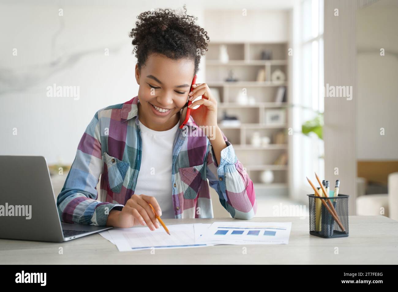 Una ragazza nera gioiosa parla al telefono mentre lavora su un notebook con documenti Foto Stock