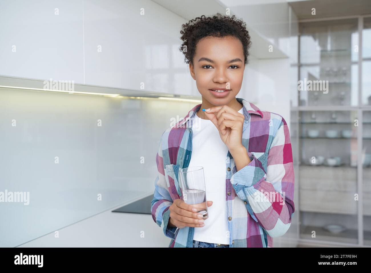 Giovane ragazza che tiene un bicchiere d'acqua in una mano e una pillola (farmaco) nell'altra, con uno sfondo luminoso e moderno della cucina Foto Stock