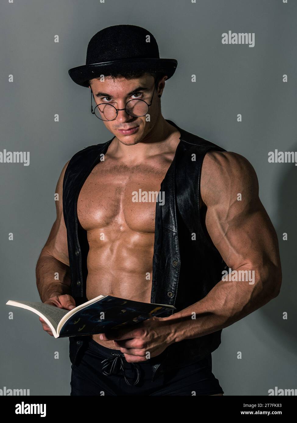 Un uomo con cappello e occhiali tiene un libro. The Intellectual Hulk: Un uomo muscoloso che abbraccia la conoscenza e la moda Foto Stock