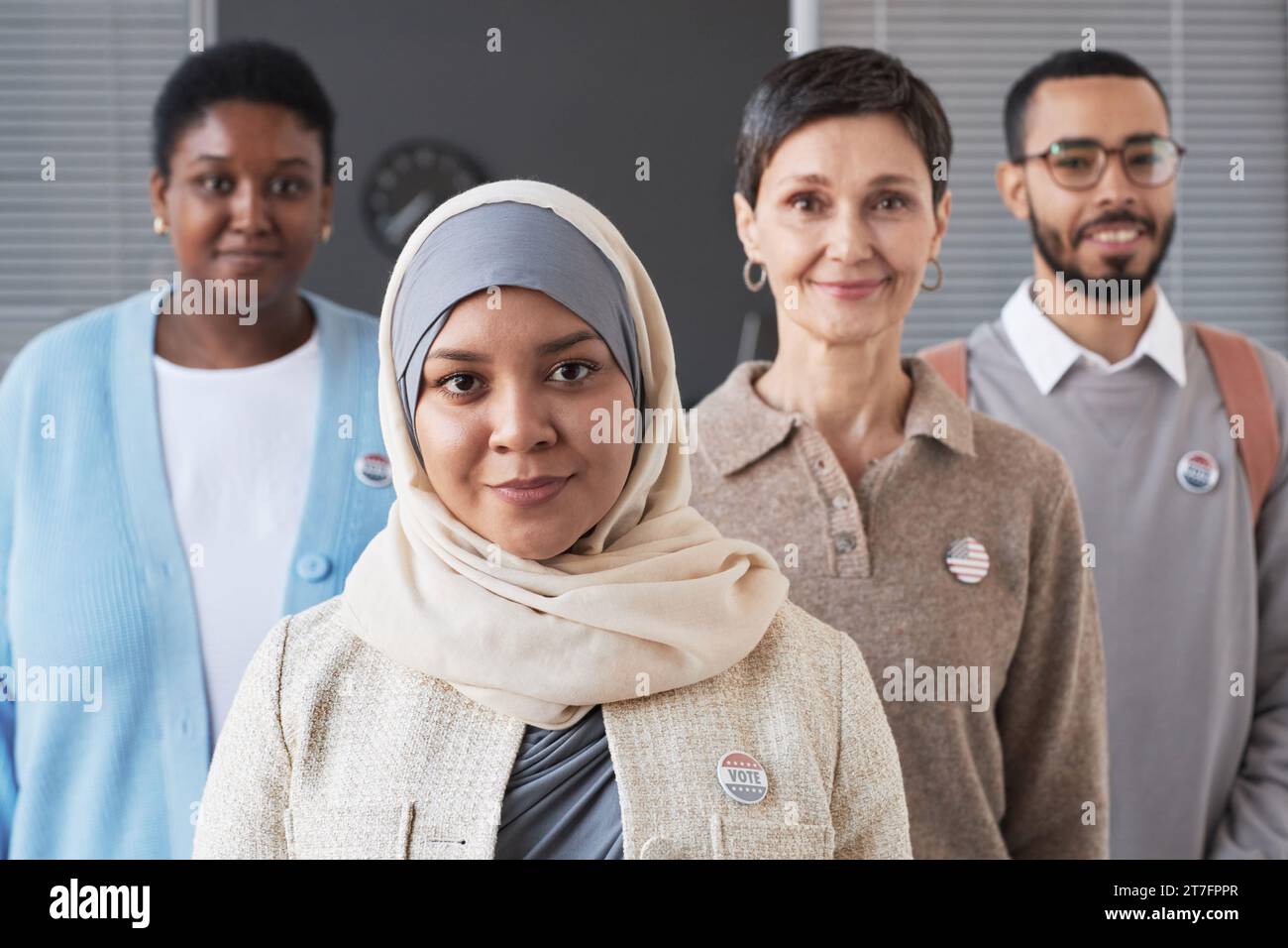 Giovane musulmana votante in hijab che guarda la macchina fotografica mentre si trova di fronte a un gruppo di persone interculturali con distintivi sul petto Foto Stock