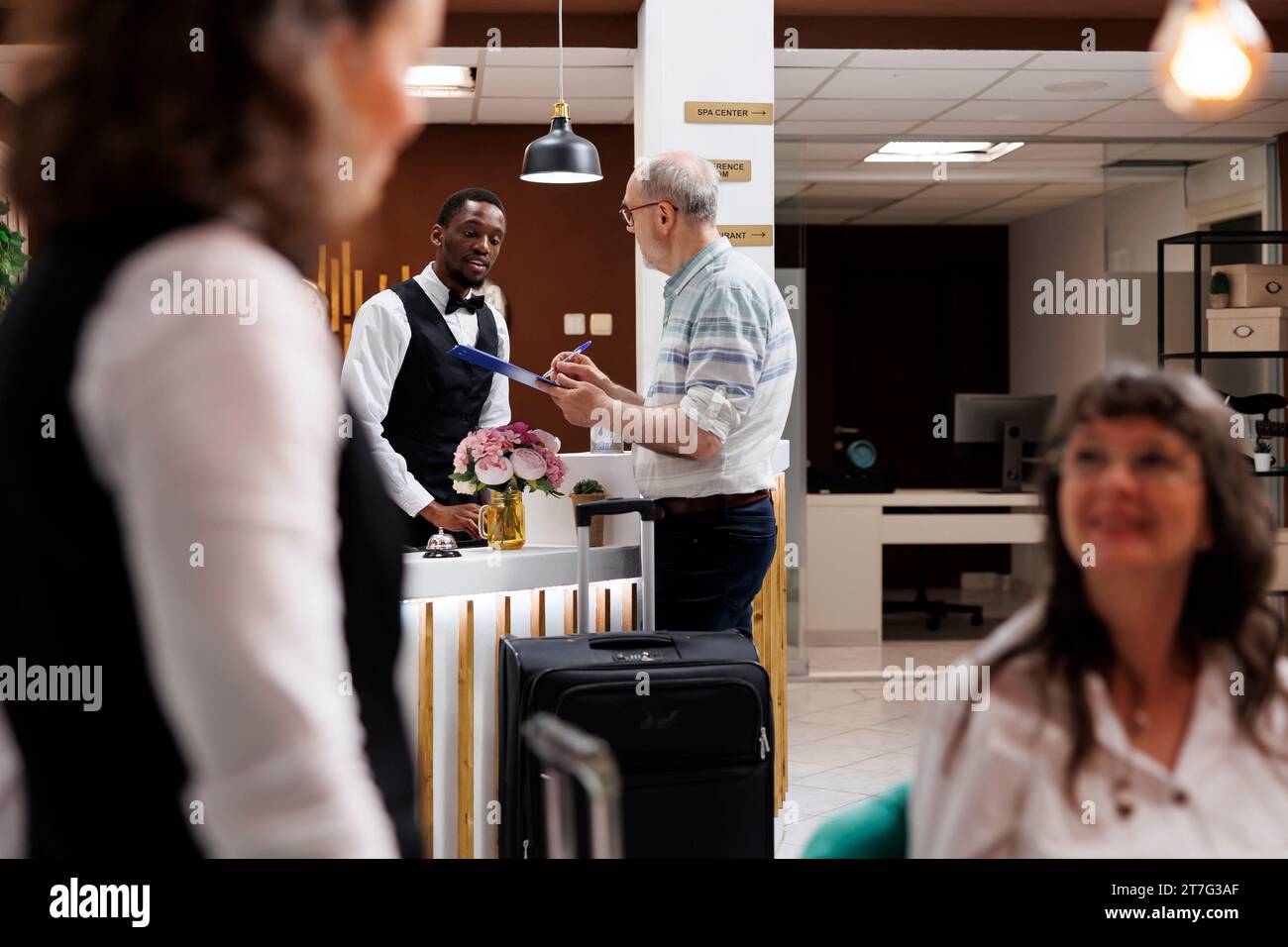 Il concierge afroamericano aiuta gli anziani in pensione nella hall dell'hotel, fornendo una procedura di check-in senza problemi. L'addetta alla reception maschile offre ai clienti anziani in pensione un cortese aiuto mentre firma il modulo di prenotazione. Foto Stock