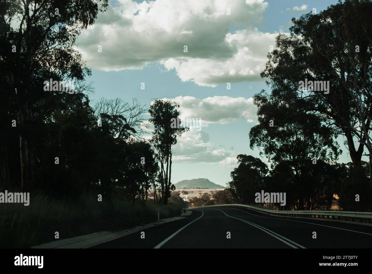 Sfreccia lungo un'autostrada attraverso una foresta nell'entroterra australiano verso un cielo spettacolare e una montagna lontana. Foto Stock