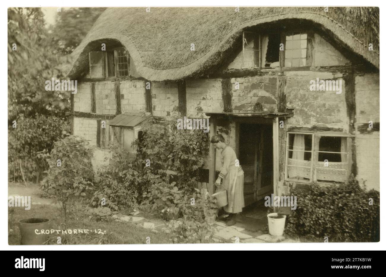 Cartolina originale del 1900 di una signora con secchio/secchio all'esterno del suo affascinante cottage inglese con tetto in paglia in legno, architettura vernacolare, risalente al periodo medievale, Cropthorne, Worcestershire. Inghilterra, Regno Unito, circa 1917 Foto Stock