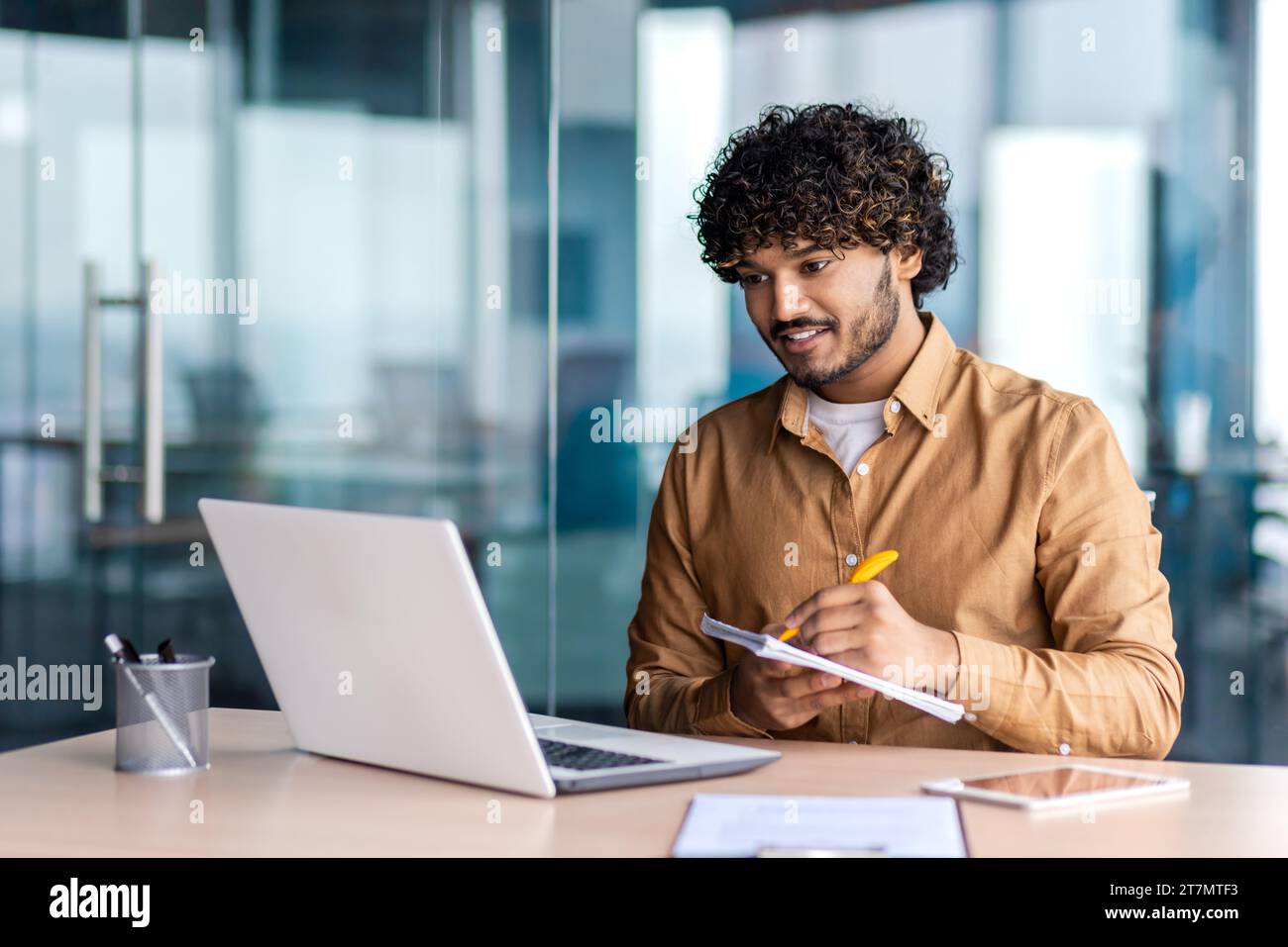 Uomo che studia online da remoto, uomo d'affari che migliora le qualifiche sul posto di lavoro all'interno dell'ufficio, scrive i dati in un notebook, guarda un corso video, soddisfatto di risultati elevati, sorride. Foto Stock