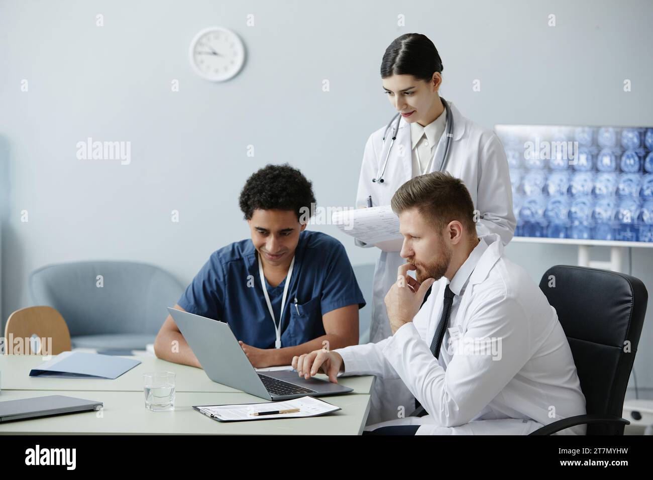Gruppo multietnico di tre giovani medici che utilizzano un computer portatile insieme in clinica con raggi X sullo sfondo, spazio di copia Foto Stock