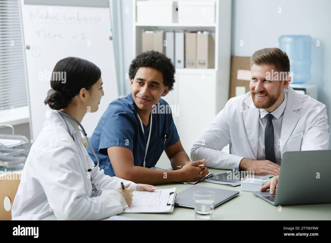 Gruppo multietnico di tre medici sorridenti che si riuniscono a tavola in clinica e discutono il caso dei pazienti Foto Stock