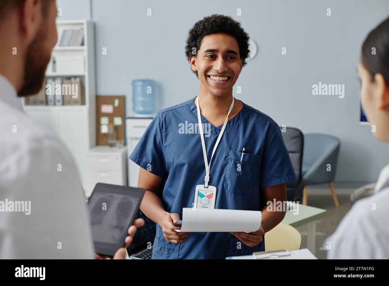 Vita su ritratto di giovane stagista medico parlando con gruppo di medici in clinica e sorridendo felicemente Foto Stock