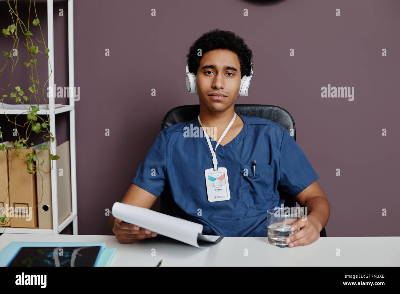 Ritratto frontale di un giovane uomo del Medio Oriente come medico maschio seduto alla reception in ufficio e indossando cuffie a cancellazione di rumore Foto Stock