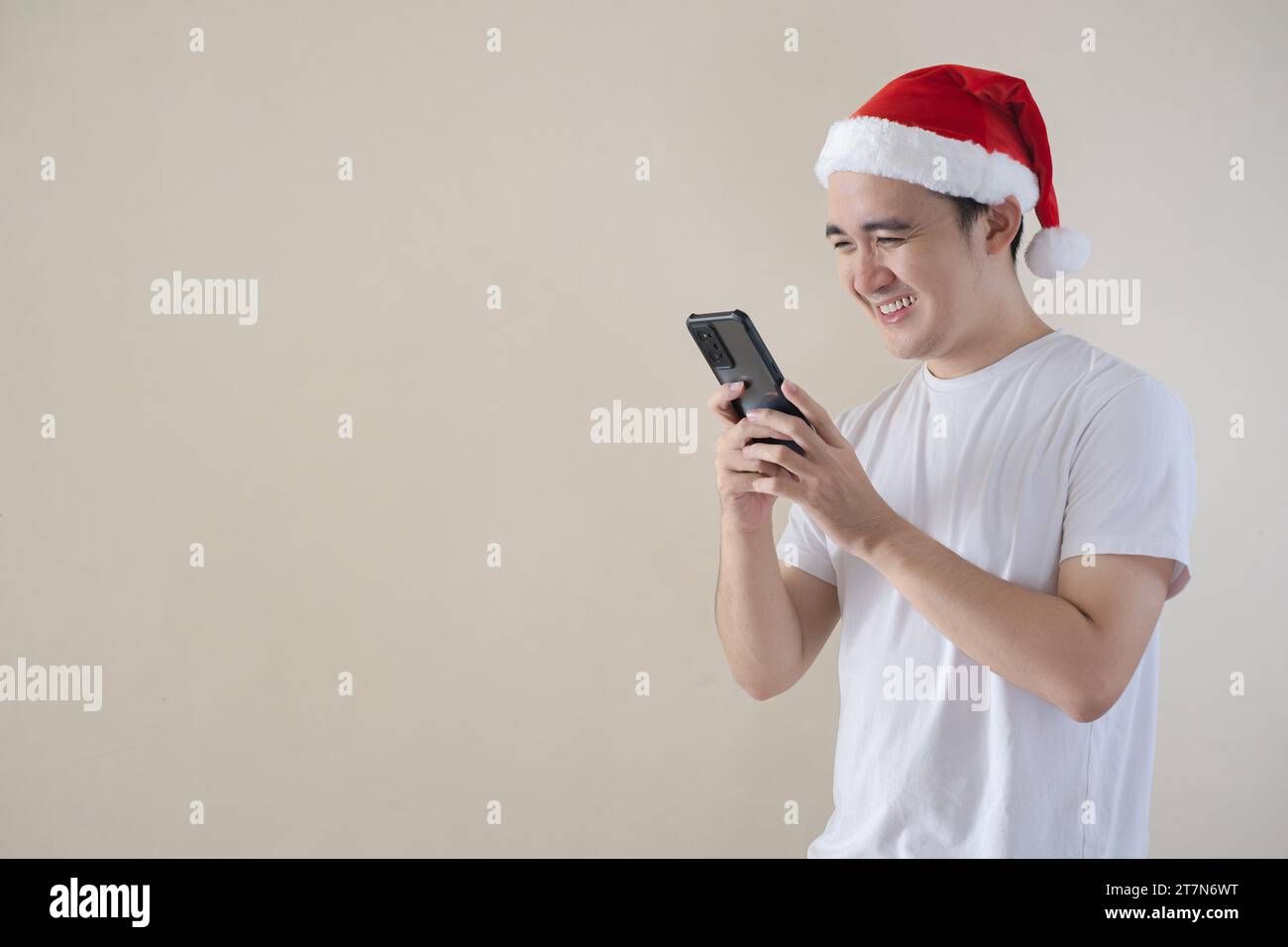 L'espressione del volto felice del giovane Babbo Natale asiatico mentre si tiene in mano e guarda uno smartphone. Sfondo beige isolato. Concetto di Natale. Foto Stock
