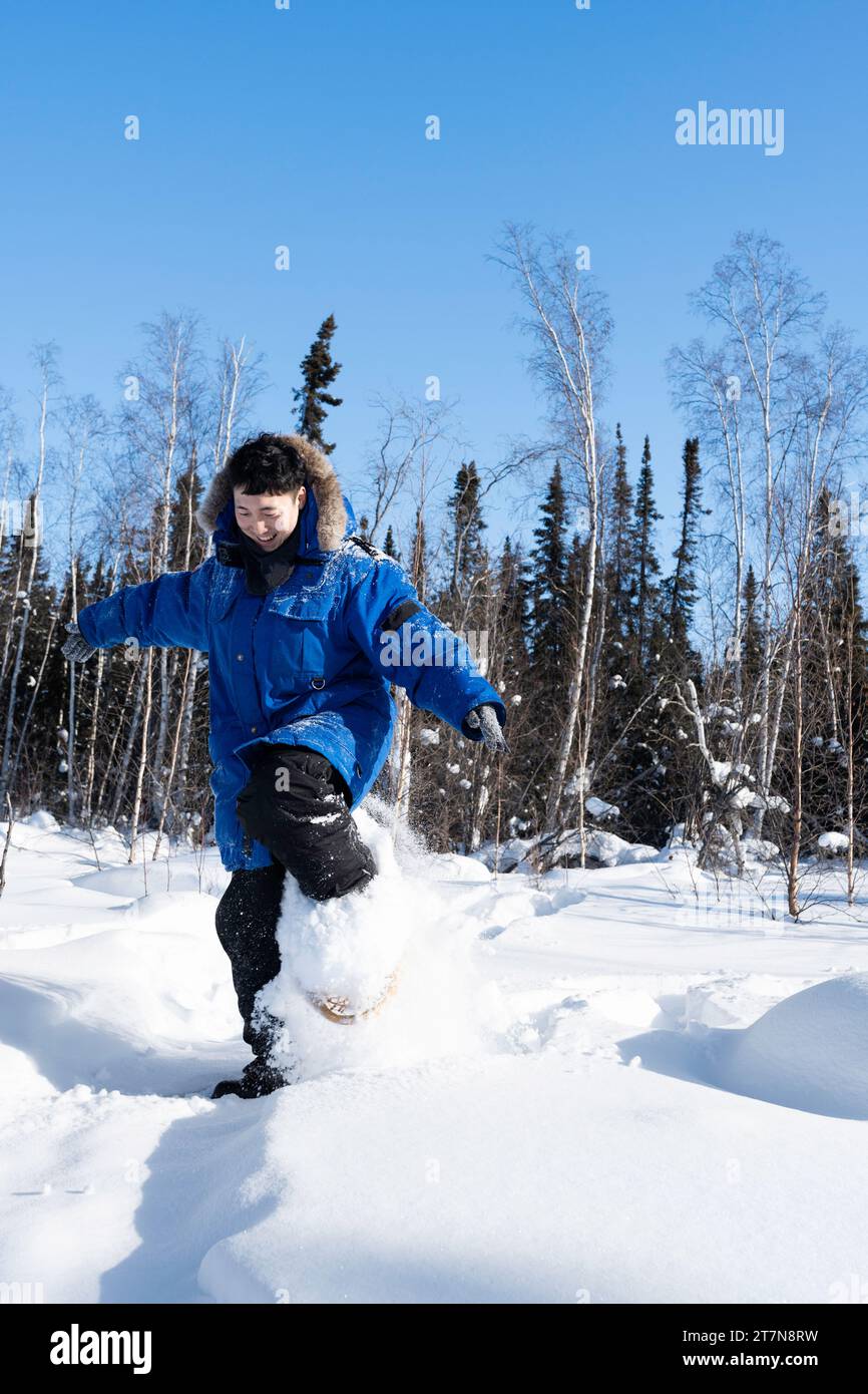 Le ciaspole di un uomo asiatico felice attraverso la neve profonda in una foresta innevata che indossa grandi racchette da neve tradizionali in legno, Yellowknife, Northwest Territories, Canada Foto Stock